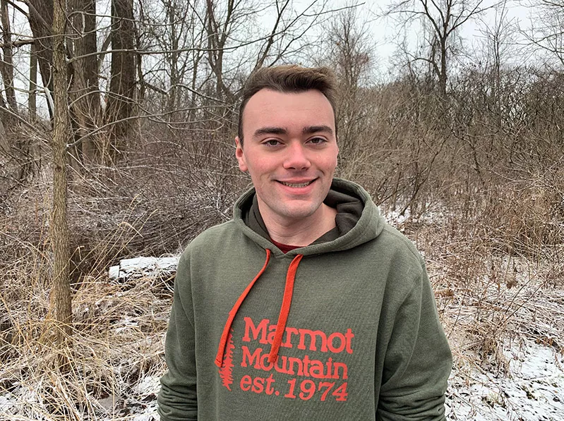 Kevin wearing a hoodie and smiling for the camera in front of some trees during winter