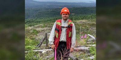 Young man in a high vis vest, hard hat and gear stands on a hill with forest in the background.