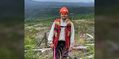 Young man in a high vis vest, hard hat and gear stands on a hill with forest in the background.