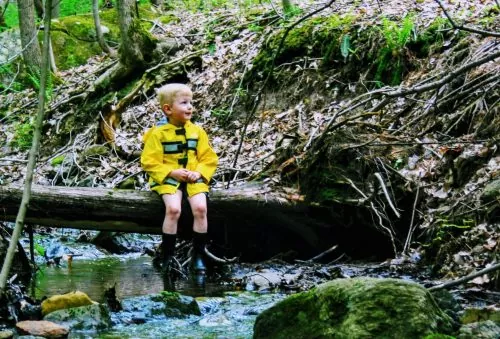 Jeune garçon en imperméable jaune assis sur un tronc d'arbre au dessus d'un ruisseau dans la forêt.