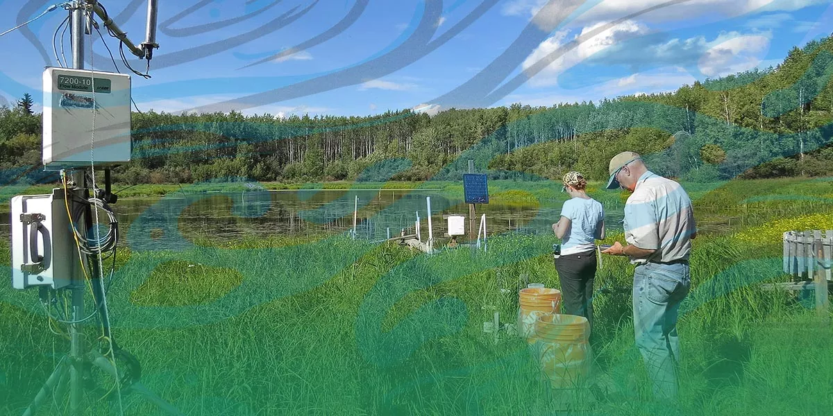 A man and woman (hydrologists) in t-shirts and jeans stand by a small lake with scientific equipment.