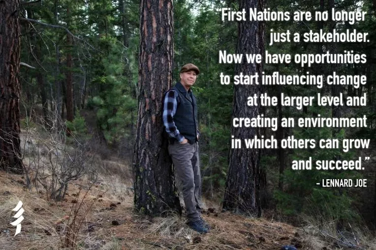Indigenous man in cap, plaid shirt and vest leans against a tree in a forest.