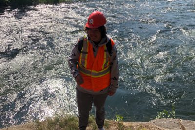 A young person stands in front of a river wearing a hard hat and a vest
