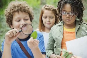 Three children learning outdoors. One examines a leaf with a magnifying glass.