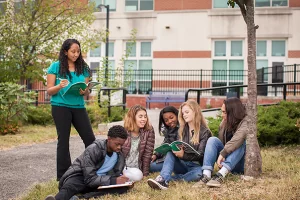 Five high school students, sitting on the ground outside, read a textbook. Their instructor stands beside them.