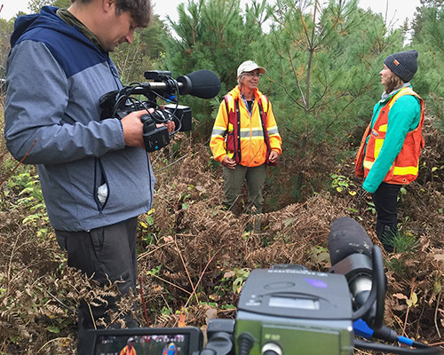 A man uses two video cameras while two women stand in vegetation wearing orange vests