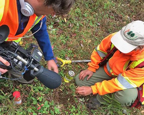 Man with video camera crouches in the dirt with woman in orange sweater and a jar of yellow liquid