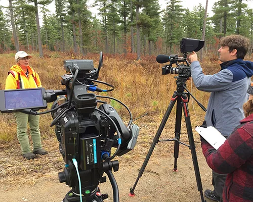 Woman in orange sweater and baseball hat talks to video cameras
