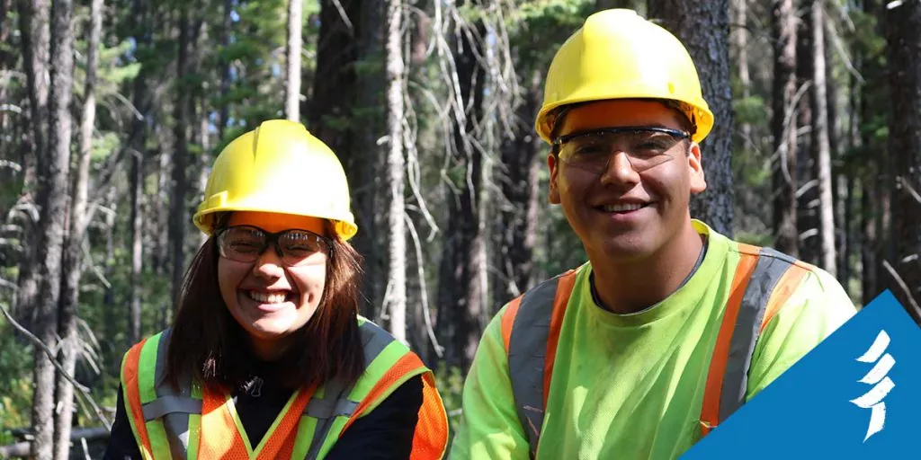 A young woman and man smiling at the camera, wearing hard hats, safety glasses, and vests