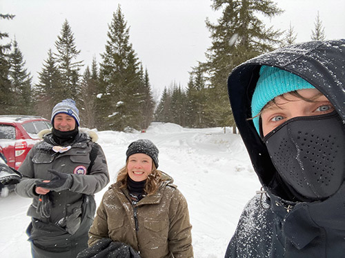 3 adults in winter parkas standing near cars with trees and snow in background