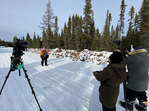 man in orange protective parka stands outdoors in front of video camera, a camera operator, and a female video host