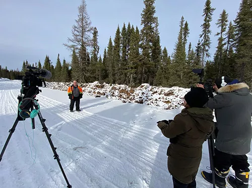 un homme en parka de protection orange se tient à l'extérieur devant une caméra vidéo, un caméraman et une animatrice vidéo