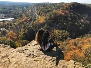 Two young women sit on top of a cliff and look down at a forest
