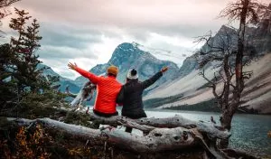Two people sit on a log and look out at the water and mountains