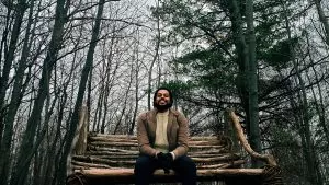 A young man sits on a bench made of branches in a forest