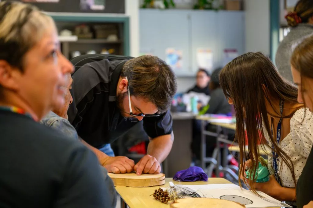 James leans over and desk and shows workshop attendees how to count rings on a tree cookie.