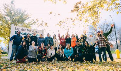 Workshop attendees and instructors stand and sit in front of the Lac des Milles Lacs Education Centre sign, throwing fallen leaves in the air.